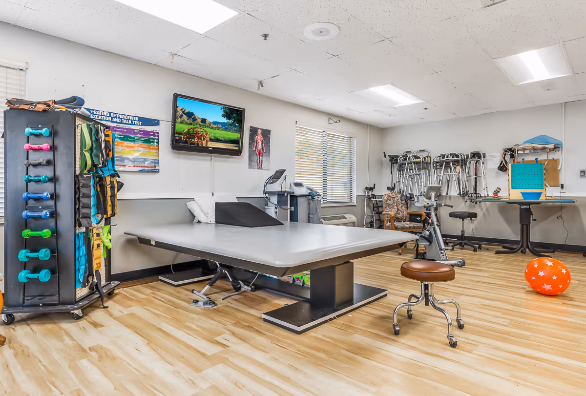 A senior facility therapy/exercise room with a large treatment table, weights, walkers, exercise bike, stools and therapy equipment.