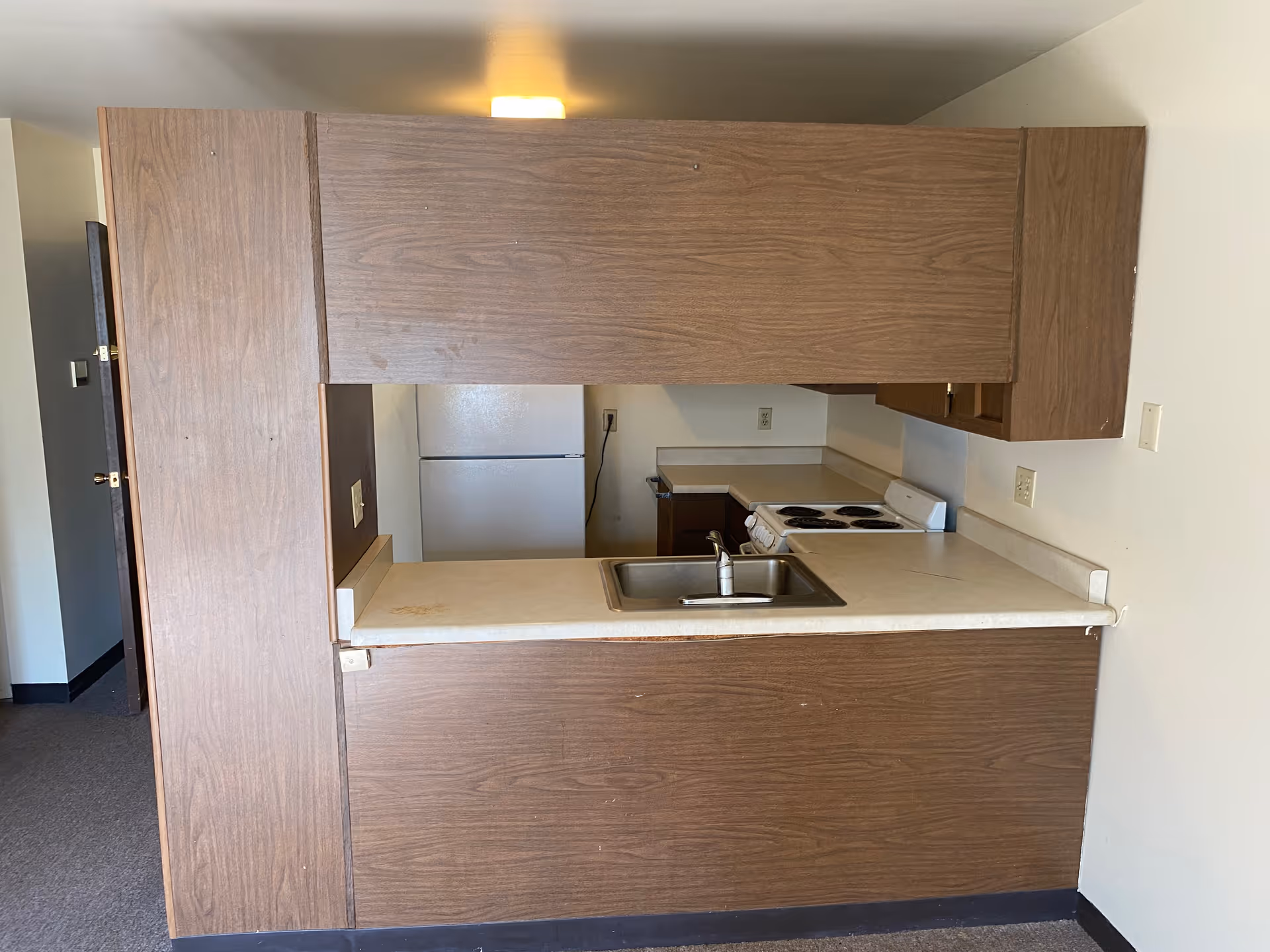 Small kitchen area with wood-paneled cabinets and a countertop with a sink. There is a white refrigerator and a white electric stove with four burners. The kitchen is open to an adjacent room with carpeted flooring and white walls.