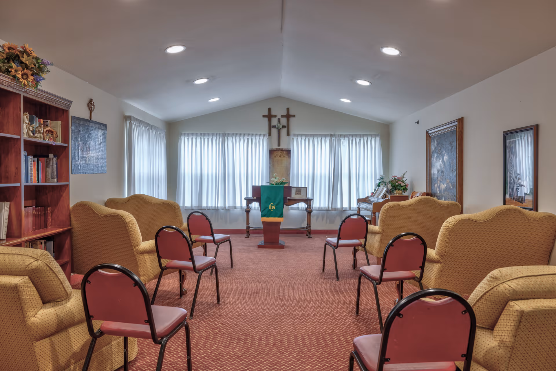 A small chapel or prayer room with beige armchairs and red chairs arranged in rows facing a wooden podium draped with a green cloth. Behind the podium are three crosses mounted on the wall and large windows with white curtains. The room has a carpeted floor, a bookshelf on the left, and framed artwork on the right wall.