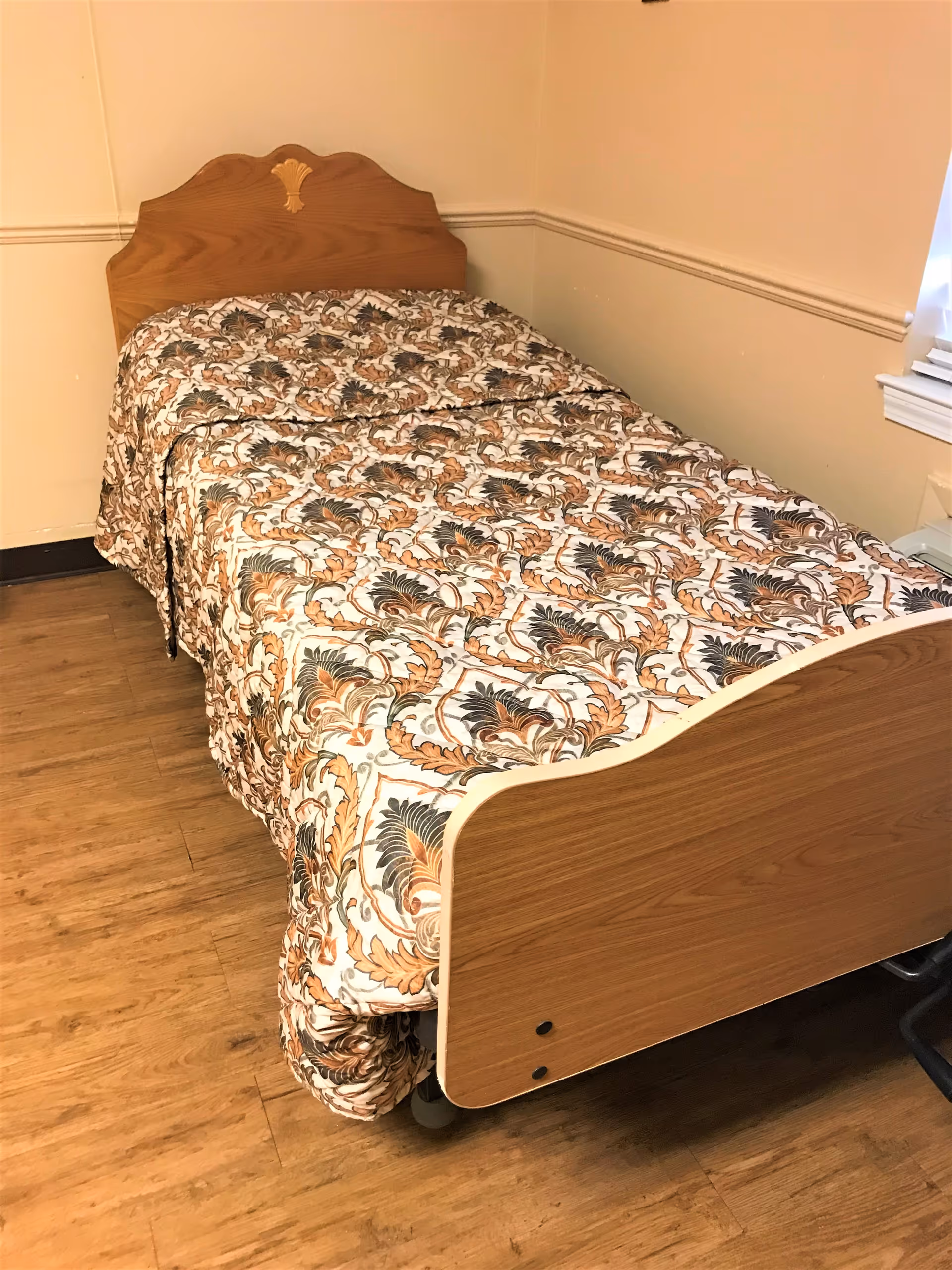 Single hospital-style bed with a floral patterned bedspread in a small room with wood flooring.