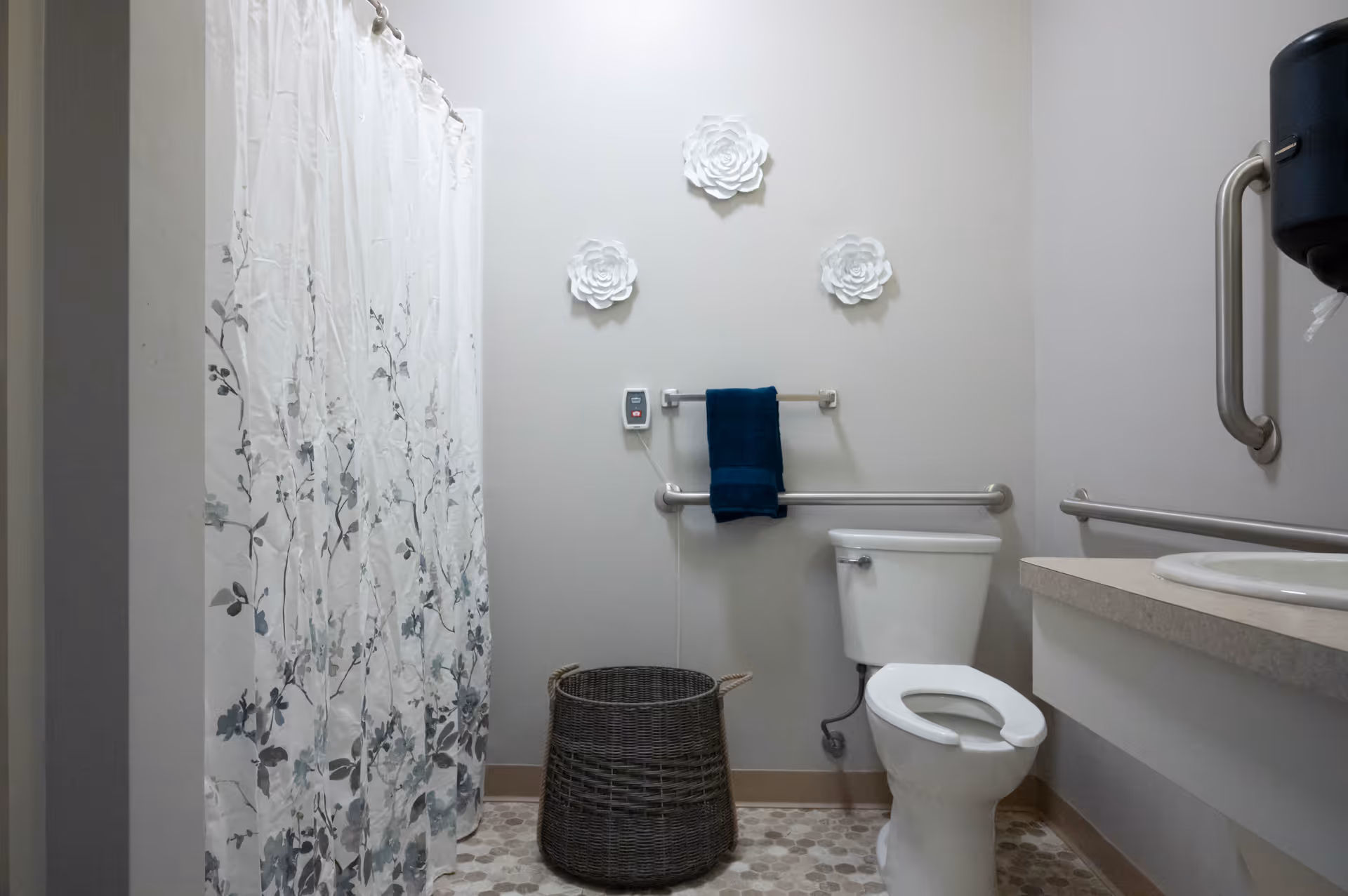 A bathroom with a white toilet, a shower with a floral curtain, a wicker laundry basket, a sink with a countertop, and three decorative white flower wall hangings. There are grab bars installed near the toilet and sink, and a dark blue towel hanging on a towel rack.