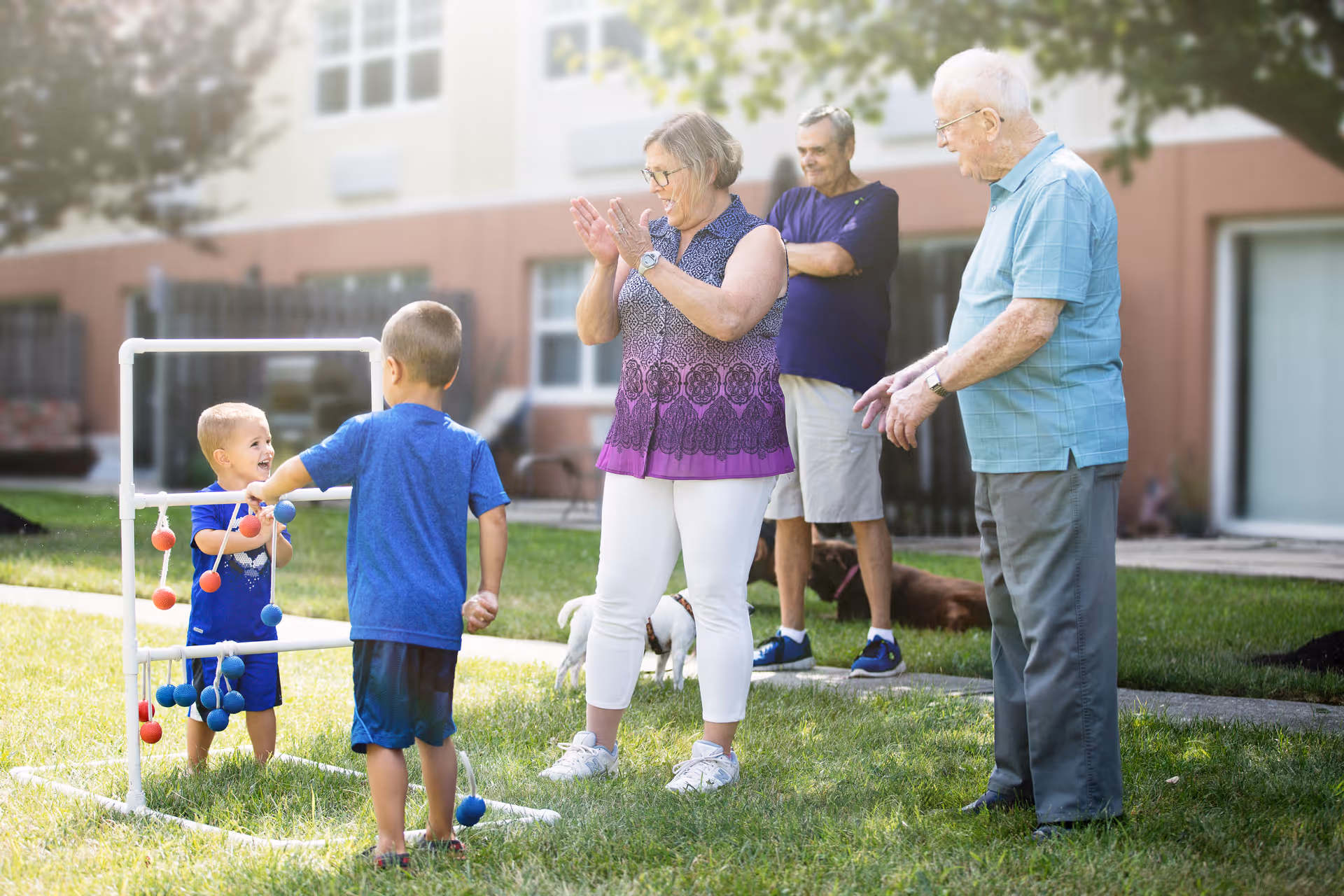 Two elderly men and one elderly woman standing on grass outside a building, watching and interacting with two young boys playing a game involving colored balls on a white frame. The elderly woman is clapping, and the group appears to be enjoying a sunny day outdoors.
