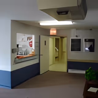 Interior hallway of a facility with beige walls and blue lower trim. There is a window on the left side showing a glimpse into another room, and an open door leading to a corridor with handrails. A small green plant is partially visible on the right side.