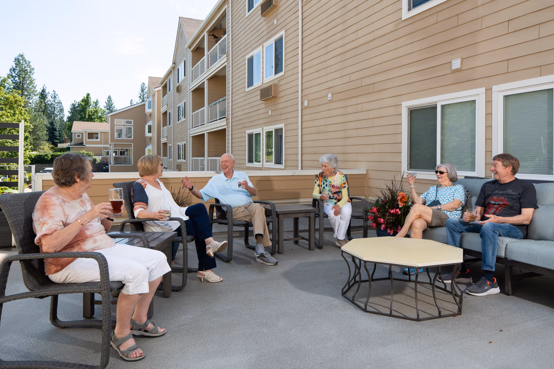 A group of six elderly people sitting on outdoor patio furniture, enjoying drinks and conversation on a sunny day outside a multi-story residential building with beige siding and white trim.