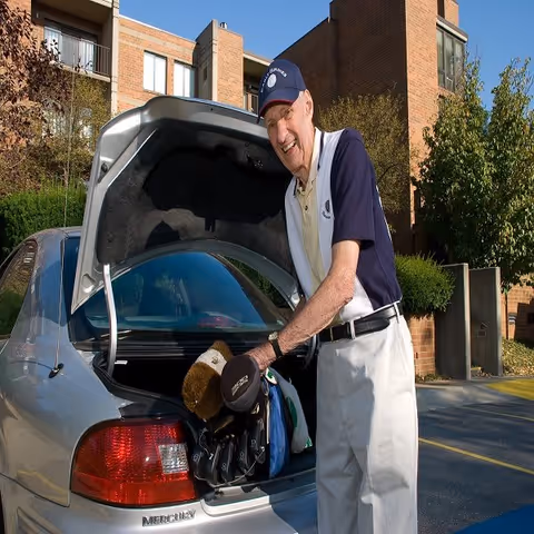 An elderly man wearing a navy and white golf shirt, white pants, and a navy cap is standing next to an open car trunk, placing or removing golf clubs from the trunk. The background shows a brick building and some greenery under a clear blue sky.