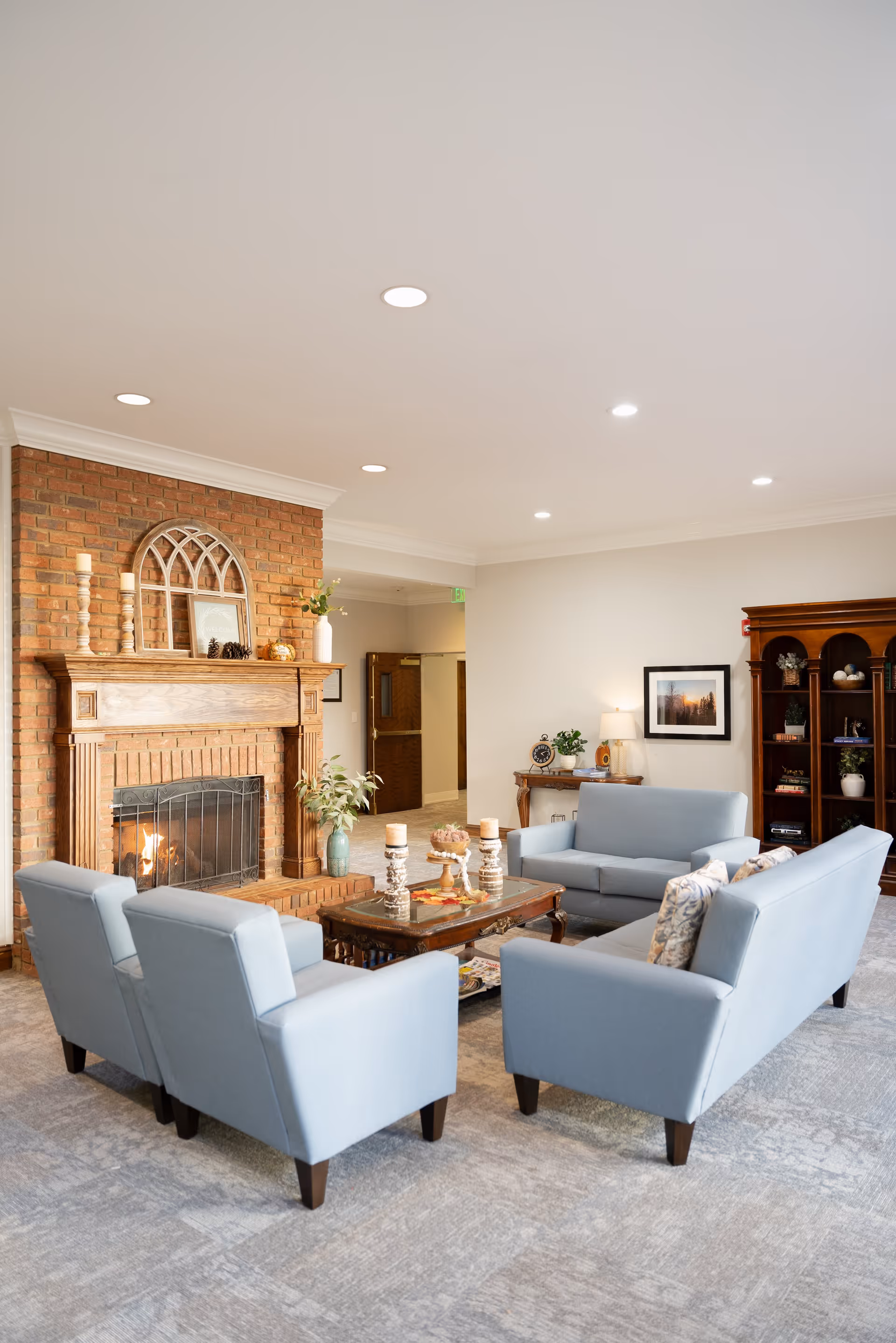 A cozy living room with light blue upholstered sofas and armchairs arranged around a wooden coffee table. The room features a brick fireplace with decorative items on the mantel, a wooden bookshelf filled with books and decor, and a side table with a lamp and framed picture on the wall. The space is well-lit with recessed ceiling lights and has a neutral color palette.