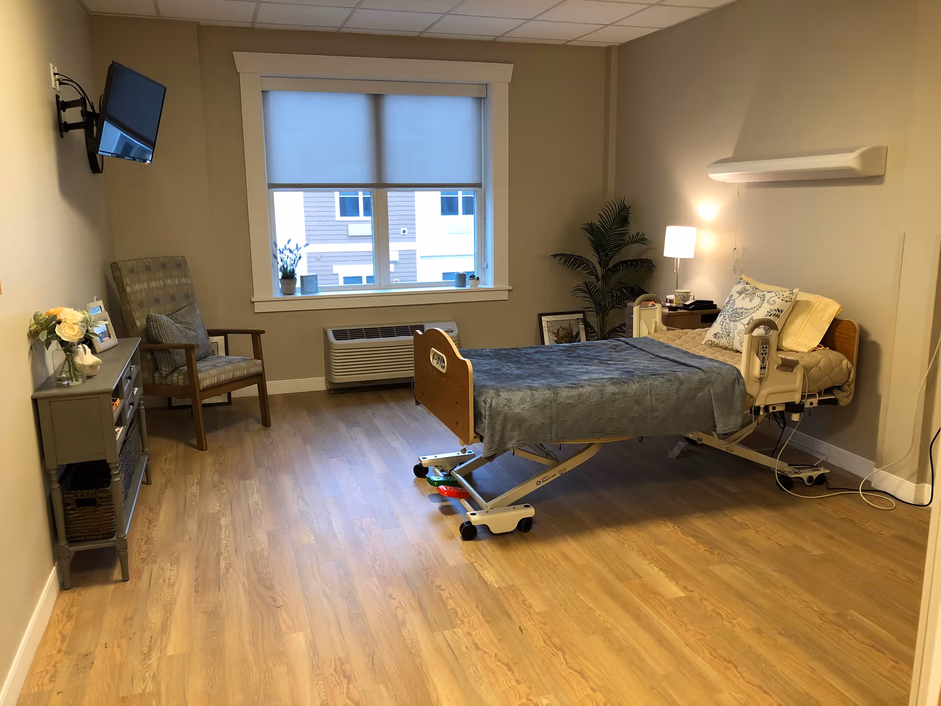 A furnished senior care bedroom with an adjustable medical bed, bedside table and lamp, armchair, wall-mounted TV and a window.