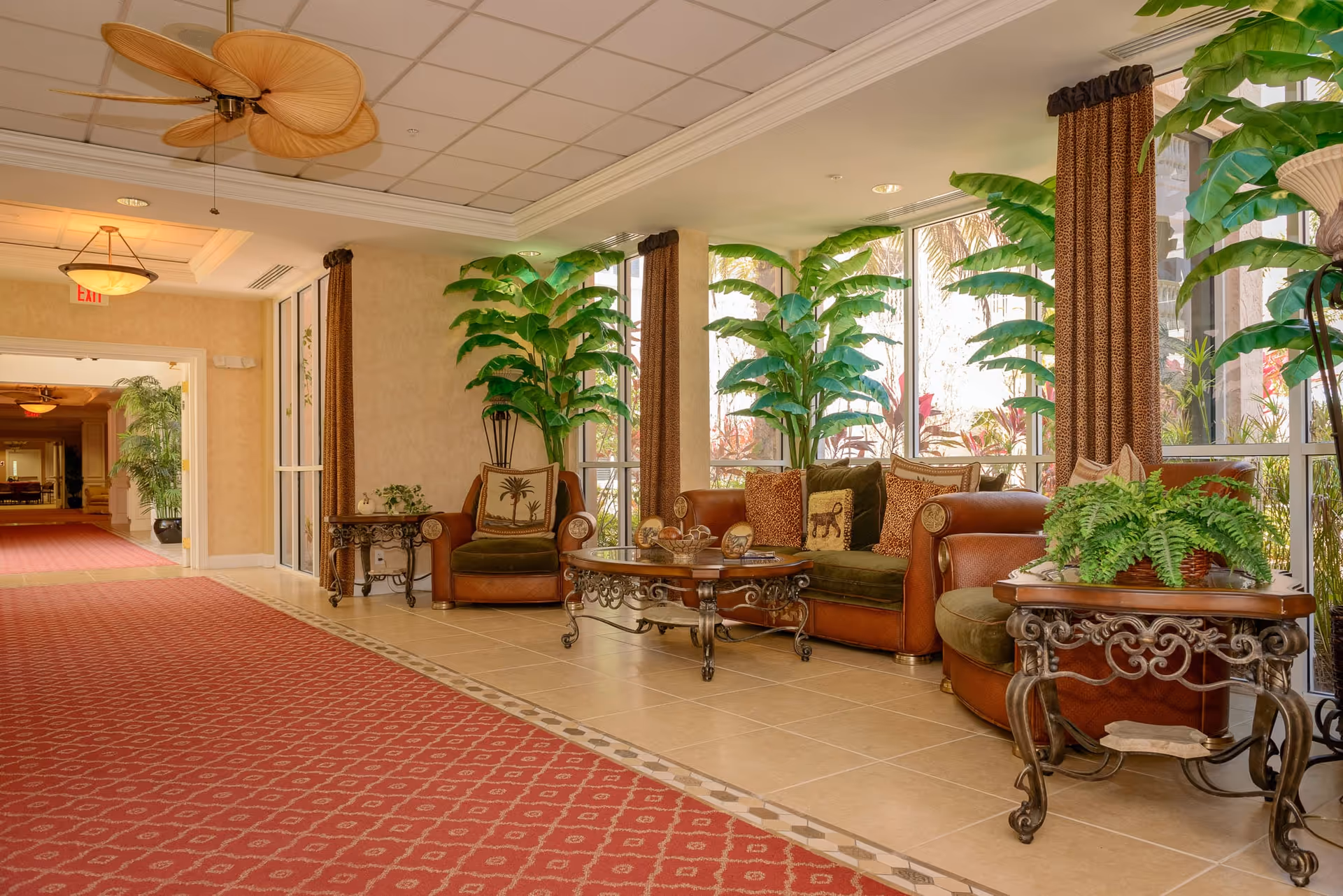 A cozy seating area in a senior living facility with leather and fabric armchairs and a sofa adorned with decorative pillows. The space features large windows letting in natural light, tall green plants, ornate side tables, a glass coffee table, and a red patterned carpet along the tiled floor. Ceiling fans and warm lighting add to the inviting atmosphere.