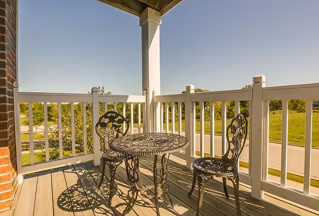 A small outdoor balcony with a round metal table and two matching metal chairs. The balcony has white railings and overlooks a green grassy area and a road under a clear blue sky.