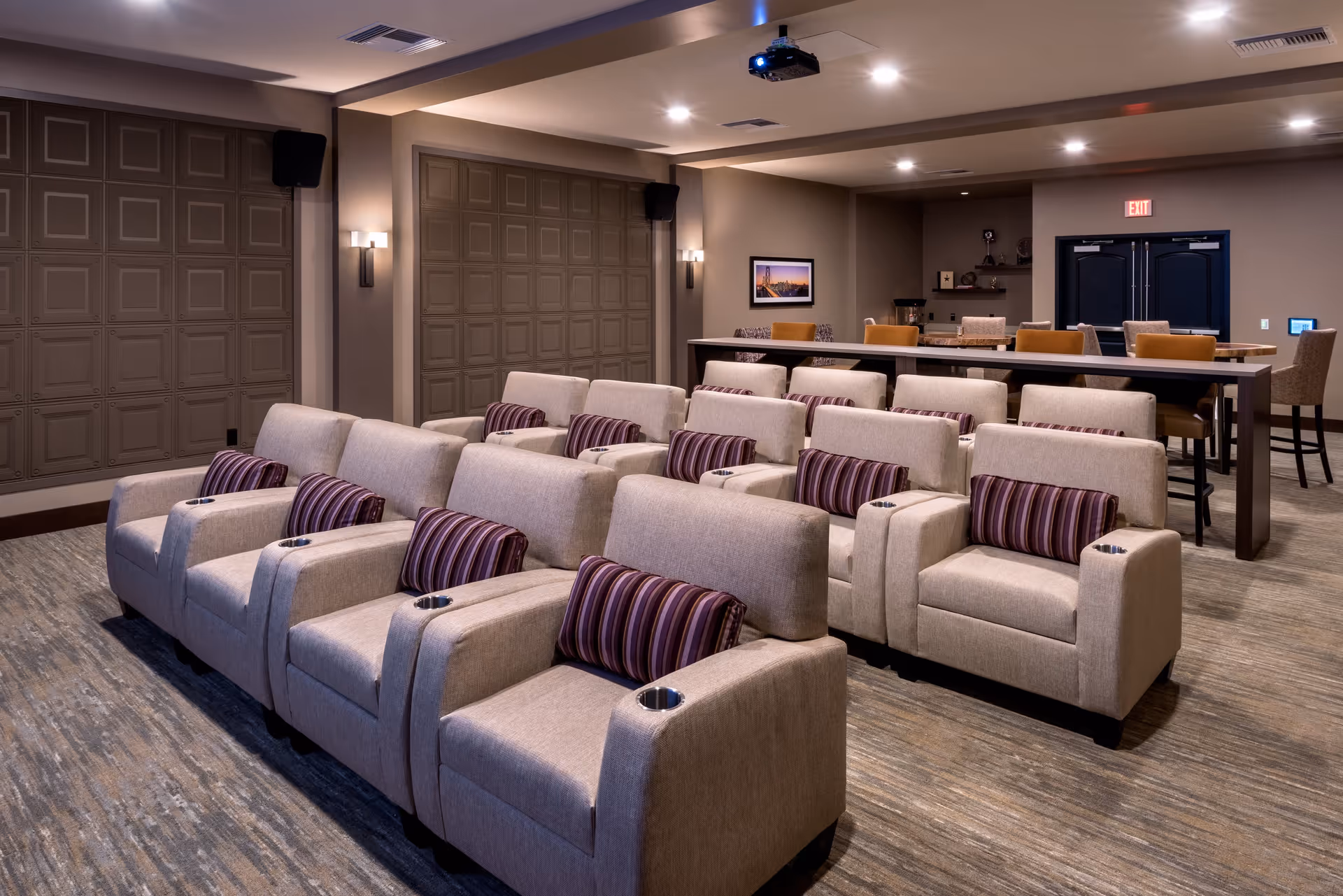 A cozy theater-style room with three rows of beige armchairs, each equipped with a striped lumbar pillow and cup holders. Behind the seating area is a long table with several chairs, and the room features soft lighting, a projector mounted on the ceiling, and decorative wall panels.