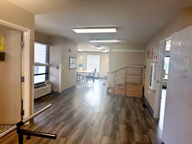 Interior view of a senior living facility hallway with wood-style flooring, beige walls, and fluorescent ceiling lights. There is a small wooden ramp with handrails on the right side, a table with chairs near a window at the end of the hallway, and an air conditioning unit below a window on the left. Doors and bulletin boards are visible along the walls.