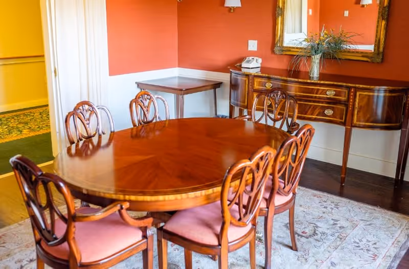 A dining room with a polished wooden oval table surrounded by six wooden chairs with pink cushions. The room has orange walls with white wainscoting, a wooden sideboard with a vase of flowers, a mirror above it, and a small wooden table in the corner. The floor is covered with a patterned rug.