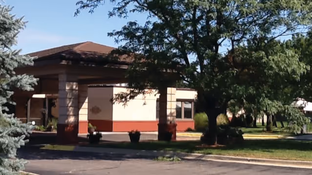 Exterior view of a single-story building with a brown roof and beige walls with red trim, surrounded by trees and greenery under a clear blue sky.