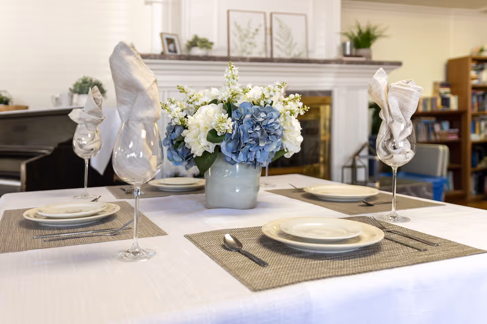 A dining table set for four with white plates, silverware, and wine glasses with folded napkins. A centerpiece of white and blue flowers in a vase is on the table. In the background, there is a fireplace with framed botanical prints and a bookshelf filled with books.