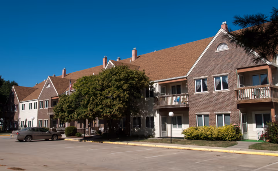 Exterior view of a two-story senior living community building with brick and siding facade, balconies, a tree, and a parking lot with a car parked.
