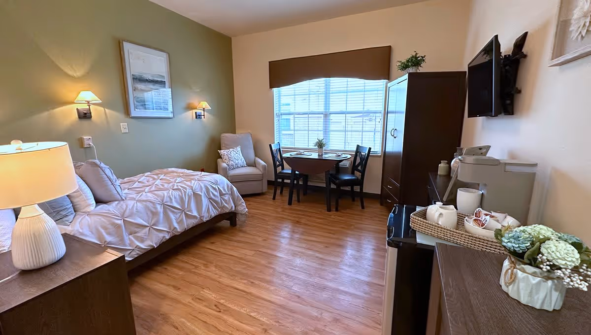 A cozy bedroom in a senior living facility featuring a single bed with a white comforter, a wooden nightstand with a lamp, a comfortable armchair with a decorative pillow, a small round dining table set for two near a large window with blinds, a dark wooden cabinet, a wall-mounted flat-screen TV, and a countertop with a coffee maker and decorative flowers.