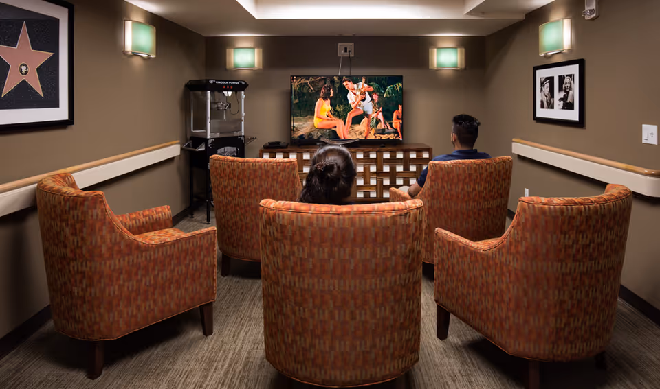 Small communal media room with upholstered armchairs facing a wall-mounted TV showing a beach scene, two people seated and a popcorn machine at the side.
