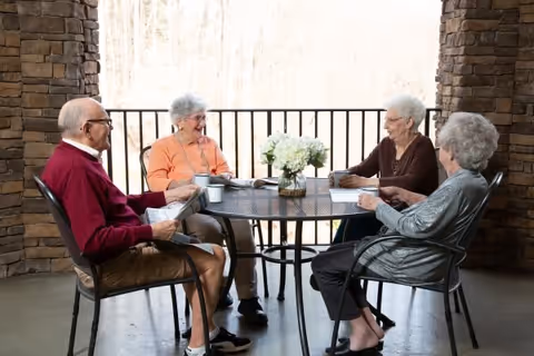 Four elderly people sitting around a round metal table on a covered patio, engaged in conversation. The table has a vase with white flowers and some cups. The background shows a railing and trees outside.