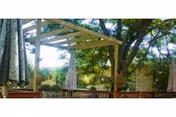 Outdoor seating area with wooden pergola, surrounded by trees and greenery, featuring umbrellas and wooden benches.