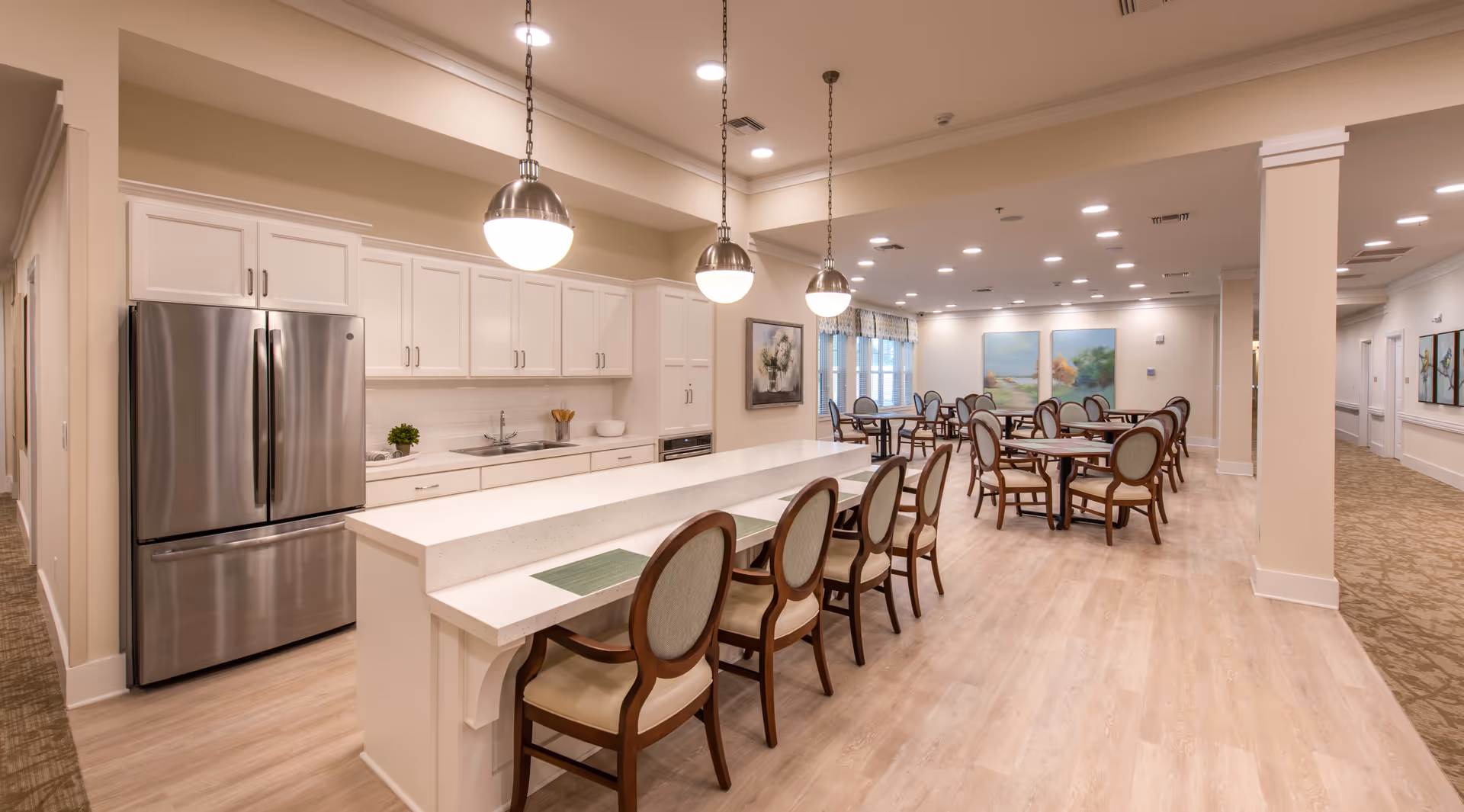 Open communal kitchen and dining area with a long island, pendant lights, stainless steel refrigerator, and multiple round dining tables and chairs.