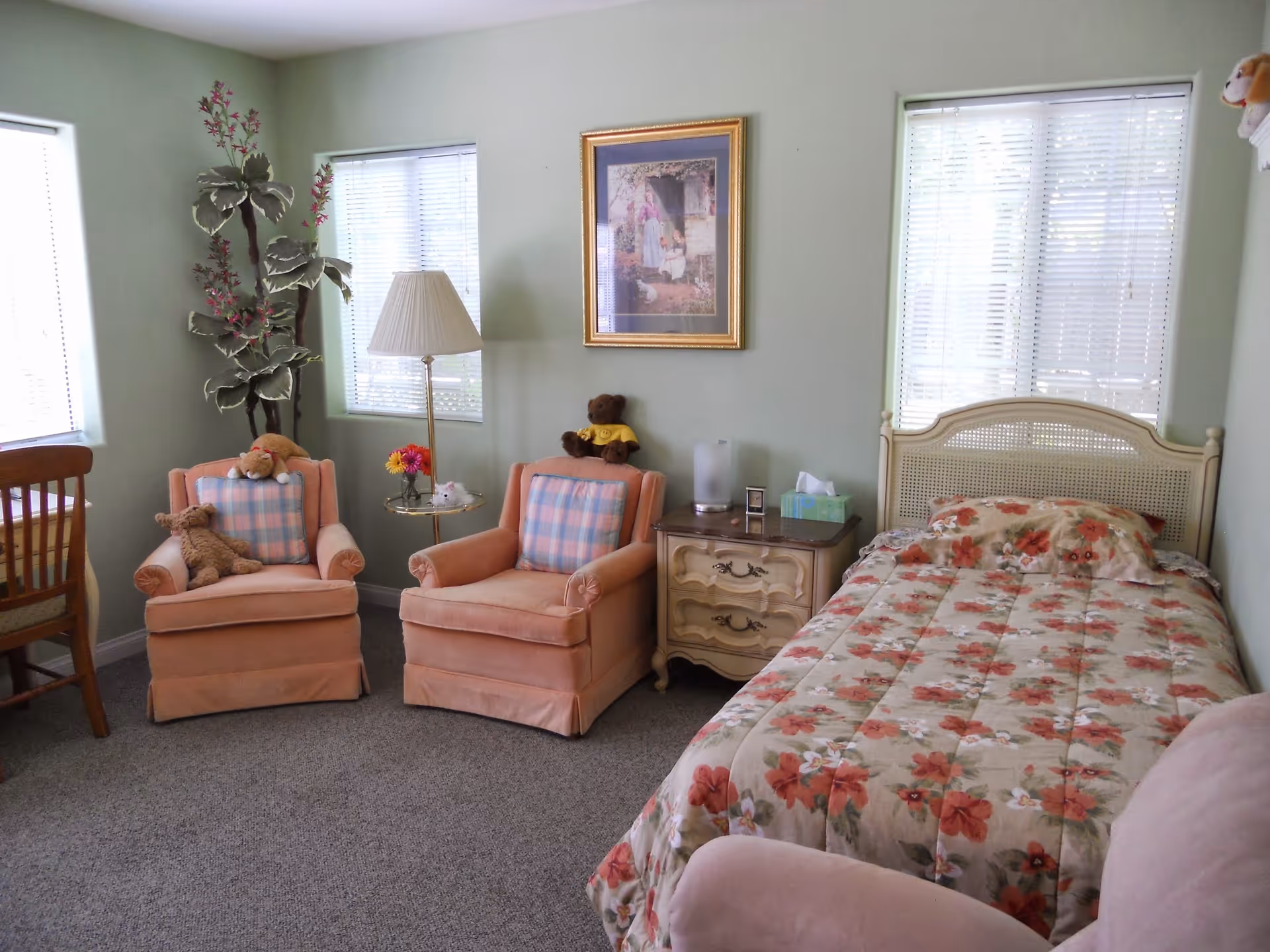 A cozy bedroom with a single bed covered in a floral bedspread, two peach-colored armchairs with plaid cushions, a wooden nightstand with a tissue box and lamp, a floor lamp, a framed picture on the wall, a wooden chair, and a tall artificial plant near two windows with blinds.