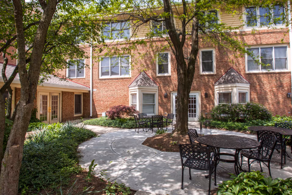 Outdoor courtyard area at The Forum at the Crossing with metal tables and chairs on a concrete patio surrounded by greenery and trees, adjacent to a brick building with multiple windows and doors.