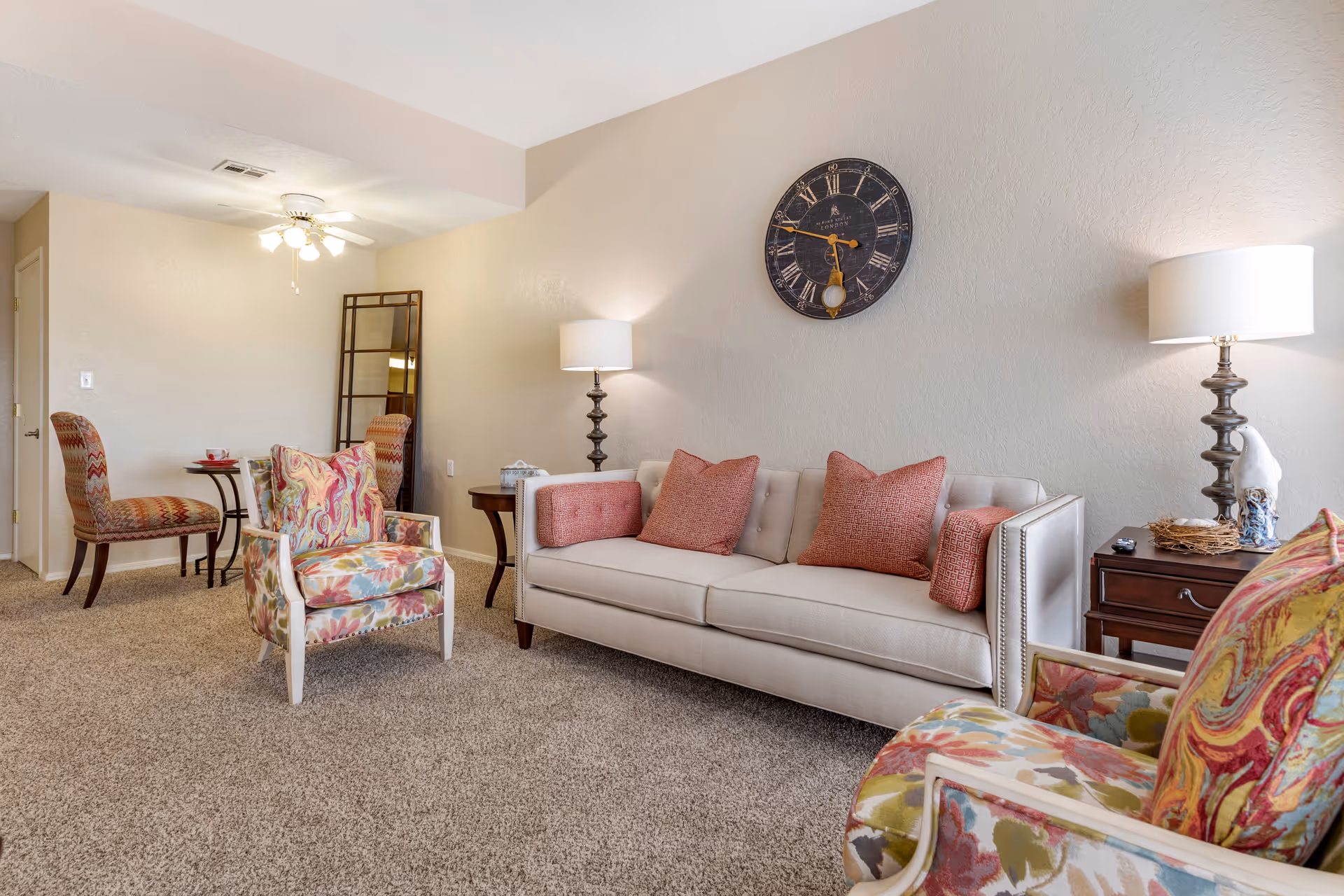 A cozy living room with a beige sofa adorned with red and patterned pillows, two floral armchairs, a round side table with a lamp, a large wall clock, and a standing mirror against a beige wall. The room has carpeted flooring and a ceiling fan with lights.