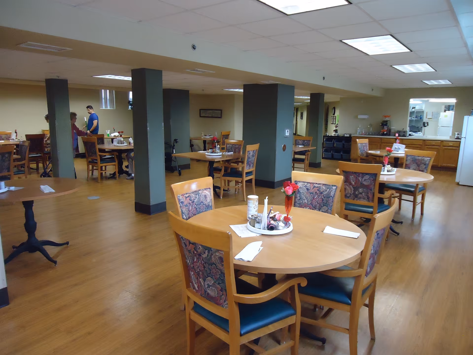 Spacious dining room with round wooden tables and chairs, floral centerpieces, and a kitchenette area in the background.