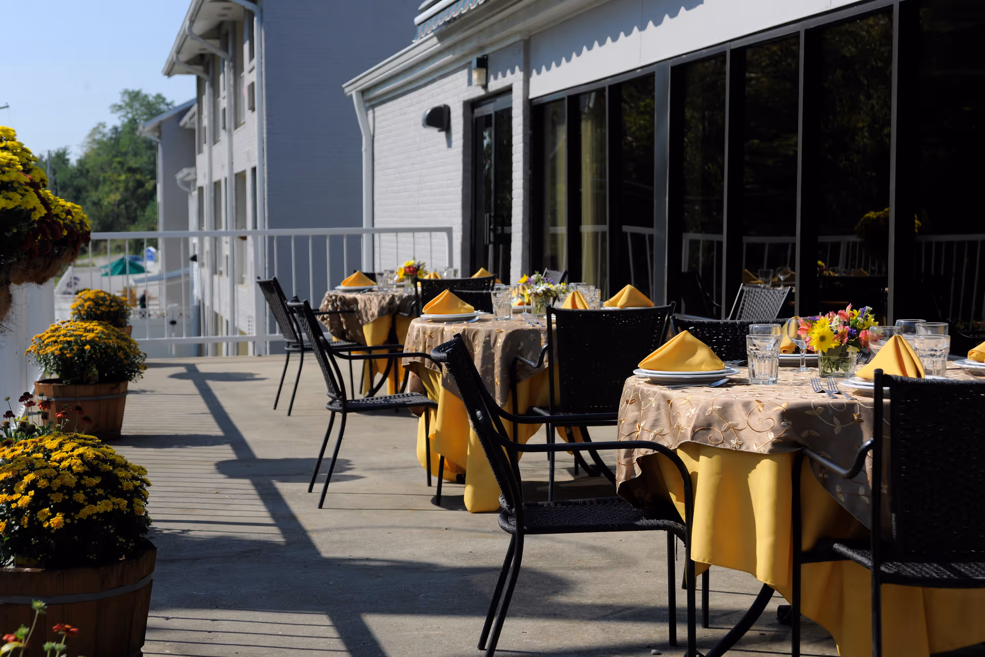 Outdoor patio area at Willow Creek Senior Living with tables covered in yellow and beige tablecloths, set with plates, glasses, silverware, and yellow folded napkins. Black chairs surround the tables, and there are flower pots with yellow flowers along the railing. The patio is adjacent to a building with large windows reflecting the outdoor scene.