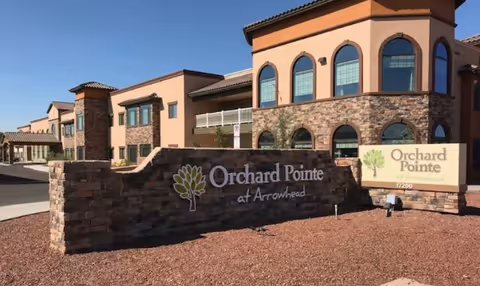 Exterior view of Orchard Pointe at Arrowhead senior living facility showing a large stone sign with the facility name and a two-story building with arched windows in the background under a clear blue sky.
