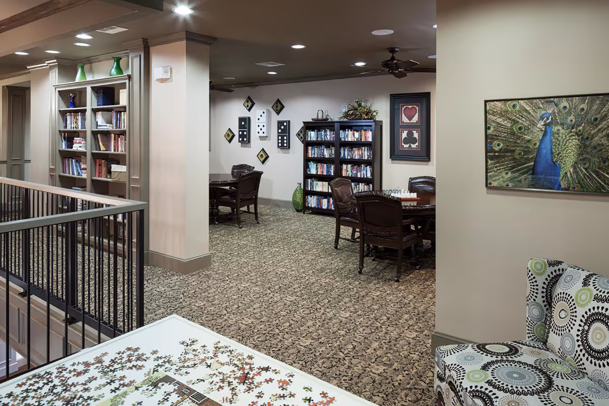 Interior view of a senior living facility common area with patterned carpet, bookshelves filled with books, several tables with chairs, a chess set on one table, decorative wall art including playing card symbols and a peacock painting, and a patterned armchair in the foreground.