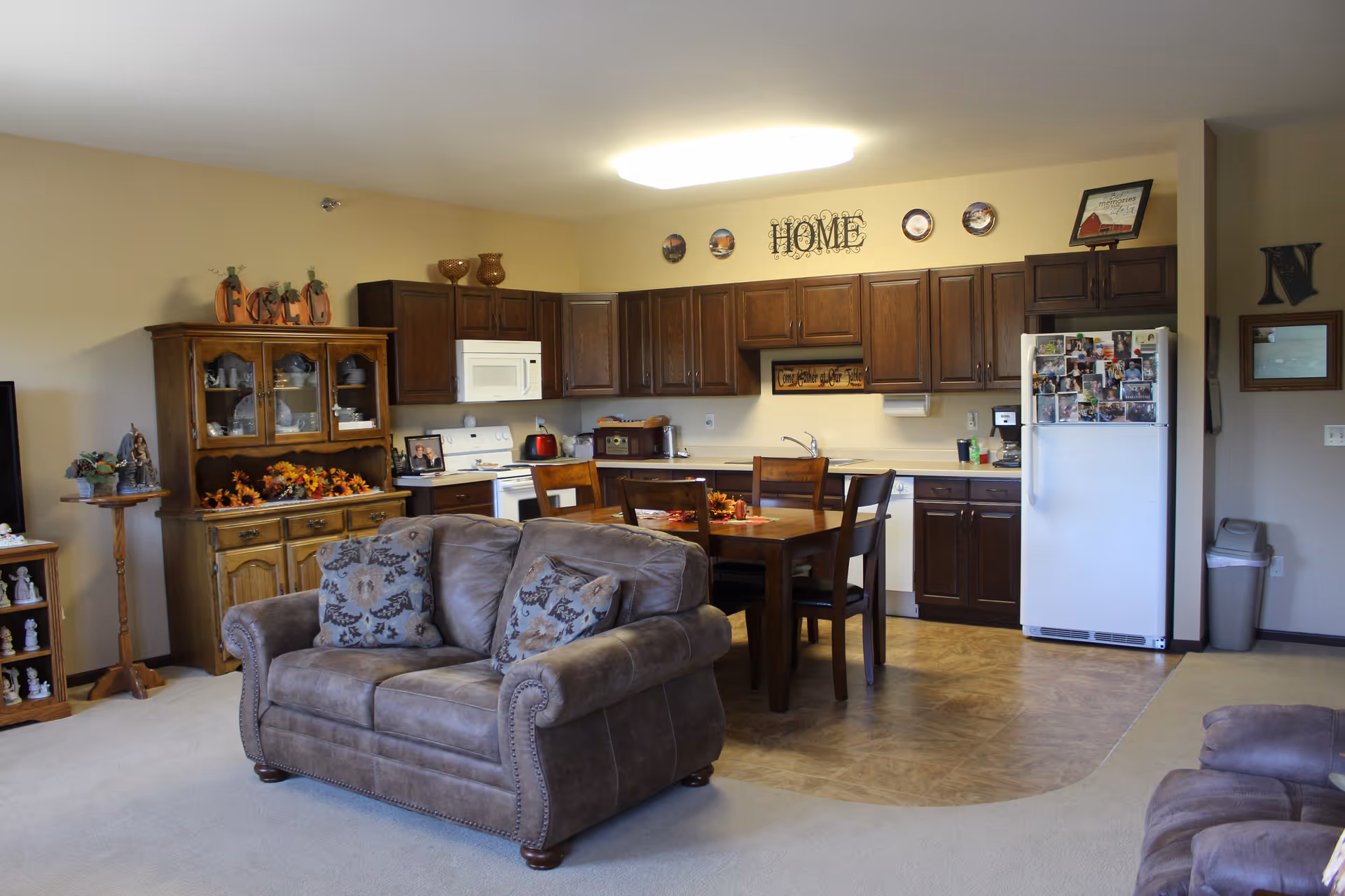 A cozy living area combined with a kitchen and dining space. The living area has a brown upholstered loveseat with patterned cushions. The kitchen features dark wooden cabinets, a white refrigerator covered with photos, a white microwave, and a stove. A wooden dining table with four chairs is placed between the kitchen and living area. Decorative items and autumn-themed decorations are placed on a wooden cabinet and shelves. The wall above the kitchen cabinets has decorative plates and a sign that reads 'HOME'.