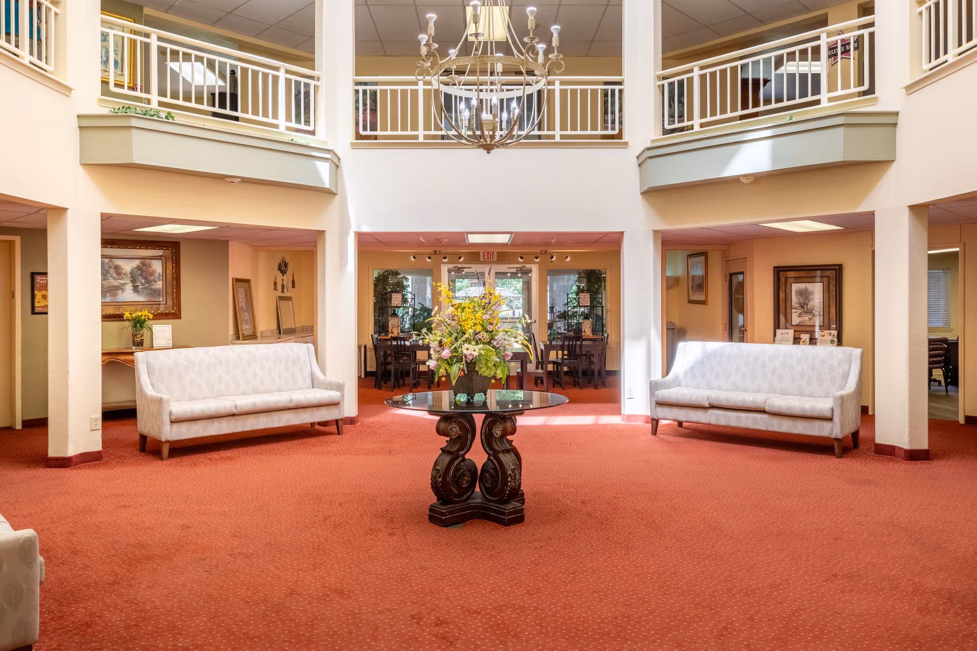 Bright senior living community lobby with a central floral table, sofas, and an upstairs balcony.