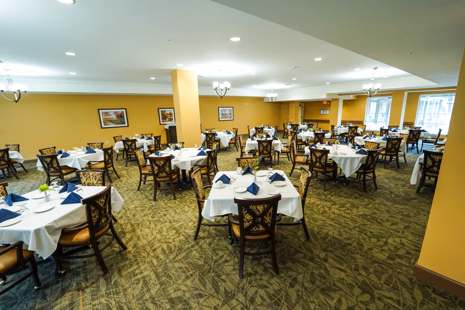 Spacious dining room with multiple round tables set with white tablecloths, navy napkins, and patterned chairs.