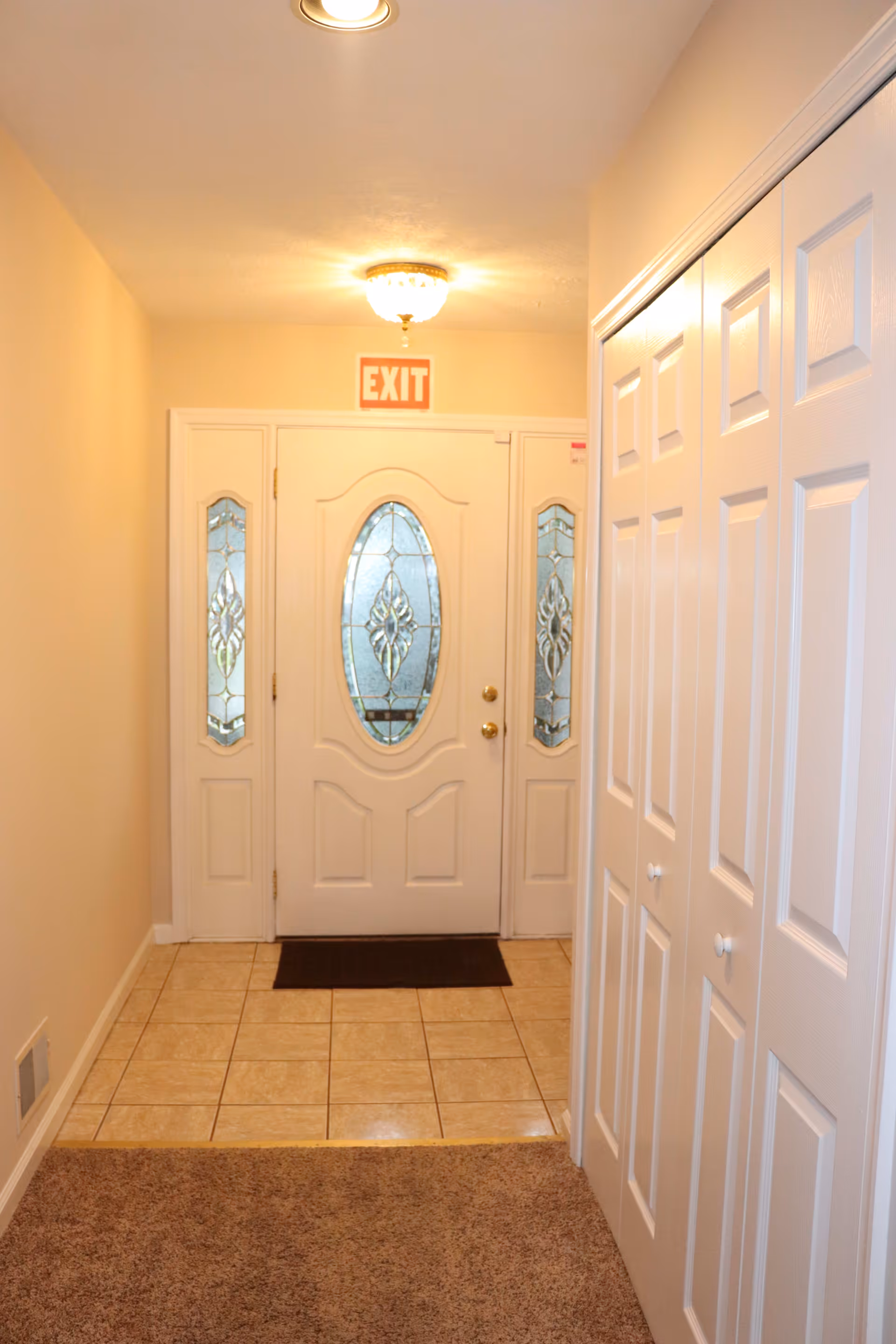Interior hallway leading to a white front door with decorative glass panels on either side. The floor near the door is tiled, transitioning to carpet in the hallway. There is an exit sign above the door and a ceiling light fixture.