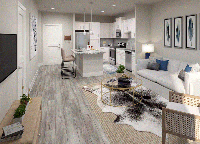 A modern open-concept living space featuring a kitchen with white cabinets, stainless steel appliances, and a granite island with two bar stools. Adjacent to the kitchen is a living room area with a white sofa, a round glass coffee table on a patterned rug, a wooden chair, and three abstract framed artworks on the wall. The floor is covered with light wood-style flooring, and there is a TV mounted on the opposite wall.