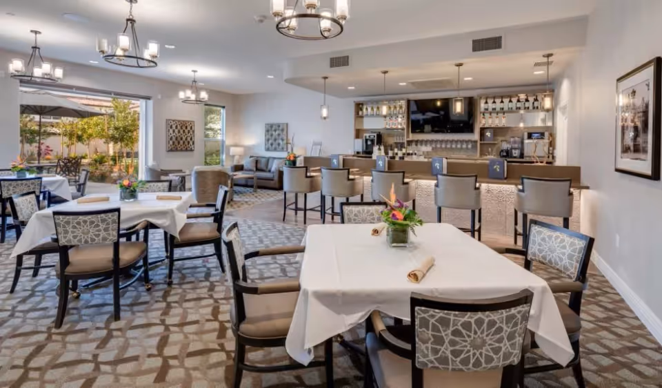 A spacious dining area with several tables covered in white tablecloths, each set with napkins and a small floral centerpiece. The room features patterned carpet, cushioned chairs, and a bar area with stools and a television mounted on the wall. Large windows and glass doors provide a view of an outdoor patio with tables and umbrellas, allowing natural light to fill the room.