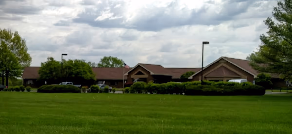 Exterior view of a single-story senior living facility building with a brown roof, surrounded by green bushes and trees, under a cloudy sky with a large grassy lawn in the foreground.