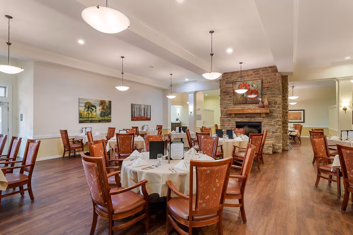 A spacious dining room with multiple round and rectangular tables covered with white tablecloths, set with glassware and napkins. The room features wooden chairs with patterned upholstery, a stone fireplace with a painting above it, and several hanging light fixtures. The walls are decorated with landscape paintings, and the floor is wooden.