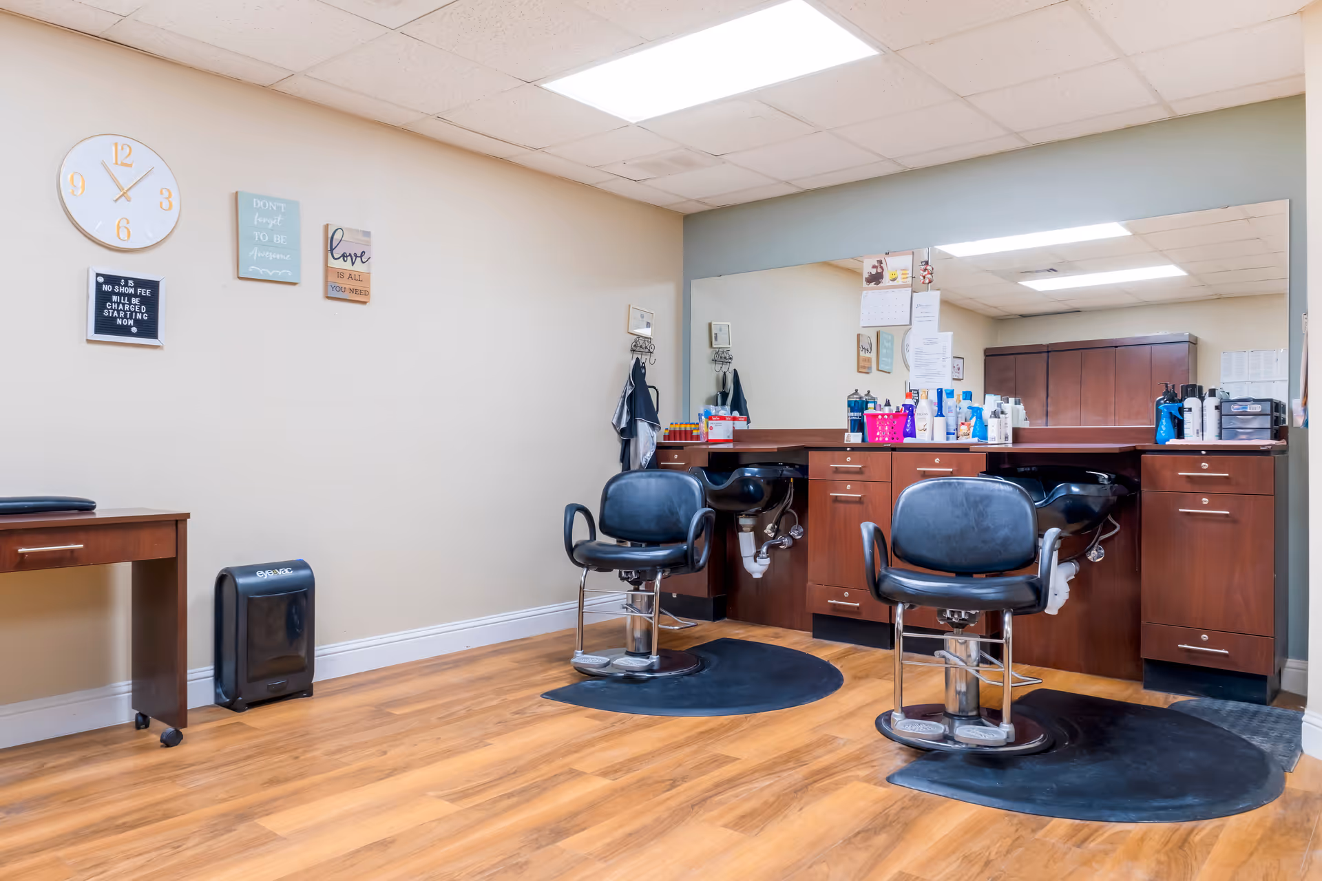 Interior view of a hair salon area in a senior living facility with two black salon chairs in front of a large mirror and wooden cabinetry. The room has light-colored walls, a wooden floor, a clock, and decorative wall signs.