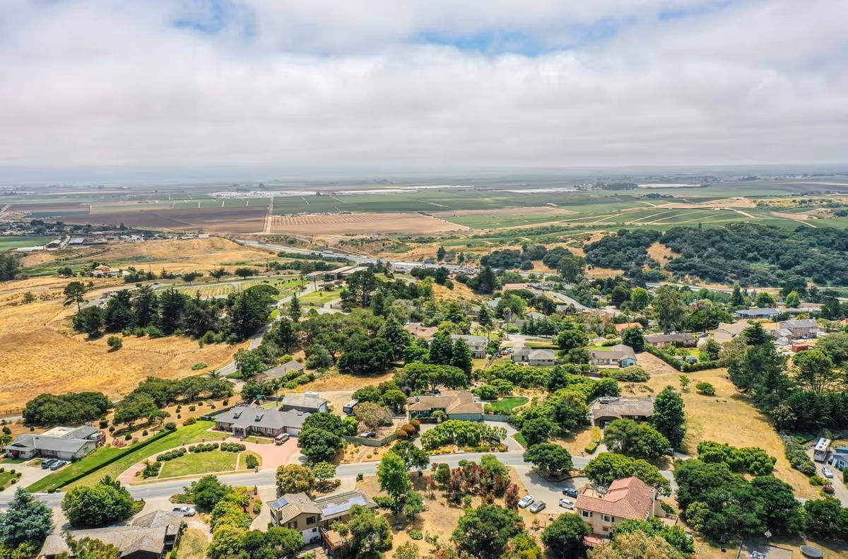Aerial view of a residential area surrounded by greenery and open fields, showing houses, trees, roads, and farmland extending into the distance under a partly cloudy sky.