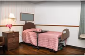 A single hospital-style bed with a pink quilted bedspread and a tray table positioned over it in a room with wooden flooring. Next to the bed is a wooden nightstand with a lamp and a vase of pink flowers. There is a framed corkboard on the wall above the bed and a window with green curtains on the right side.