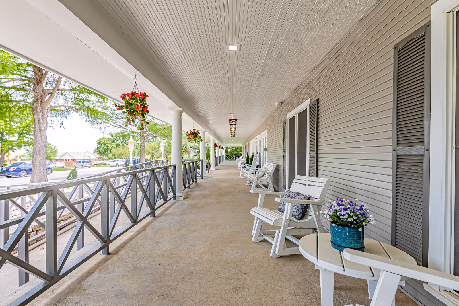 A long covered porch with white wooden rocking chairs and small tables, some with potted flowers. The porch has a beige floor, white ceiling with recessed lighting, and gray siding with shuttered windows. Hanging flower baskets are suspended from the ceiling, and trees and parked cars are visible outside the porch railing.