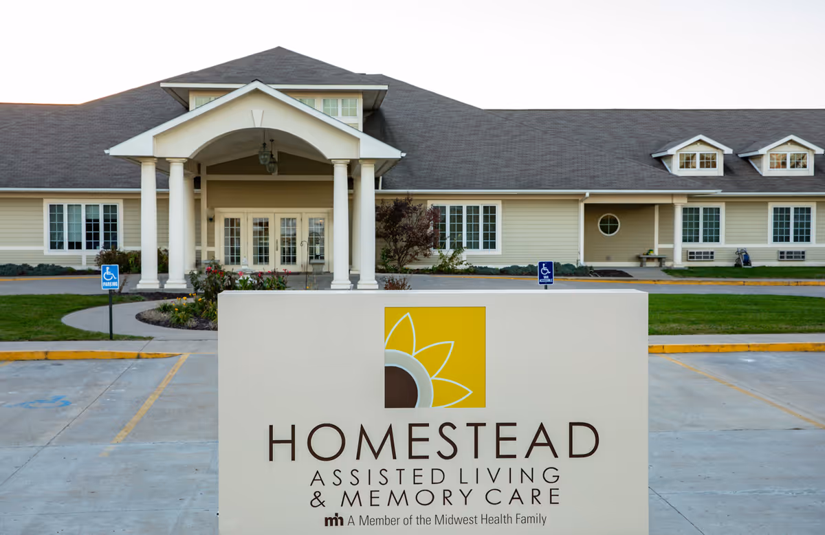 Exterior front view of Homestead Assisted Living & Memory Care facility with a large sign in the foreground displaying the facility's name and logo. The building has a covered entrance with columns, multiple windows, and a well-maintained parking area with handicap parking spaces.