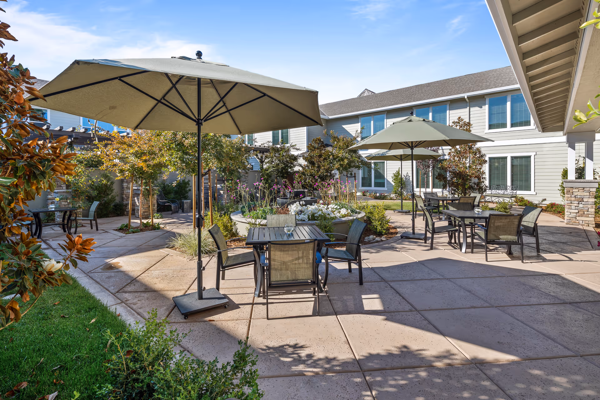 Sunny courtyard patio with tables, chairs, large umbrellas, planted flower beds, and a two-story building in the background.