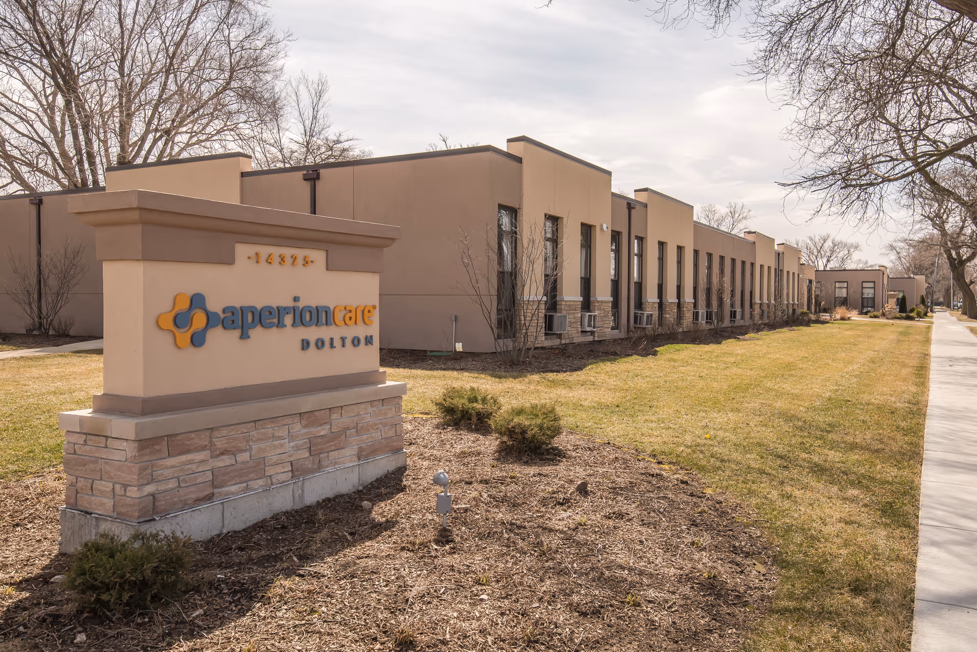 Exterior view of Aperion Care Dolton building with a sign in the foreground displaying the facility name. The building is a single-story structure with multiple windows and a beige facade. There is a grassy area and sidewalk alongside the building, with leafless trees in the background under a cloudy sky.