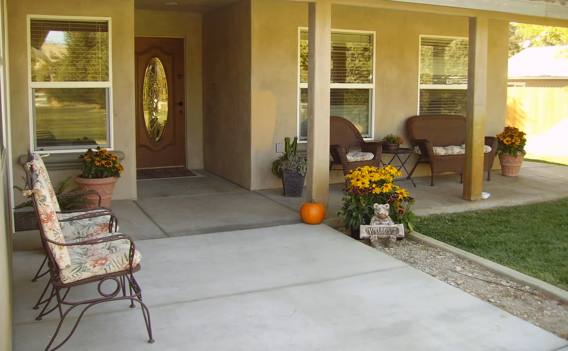 Covered front porch with seating, potted flowers, a pumpkin and a decorative welcome sign by a brown entry door.