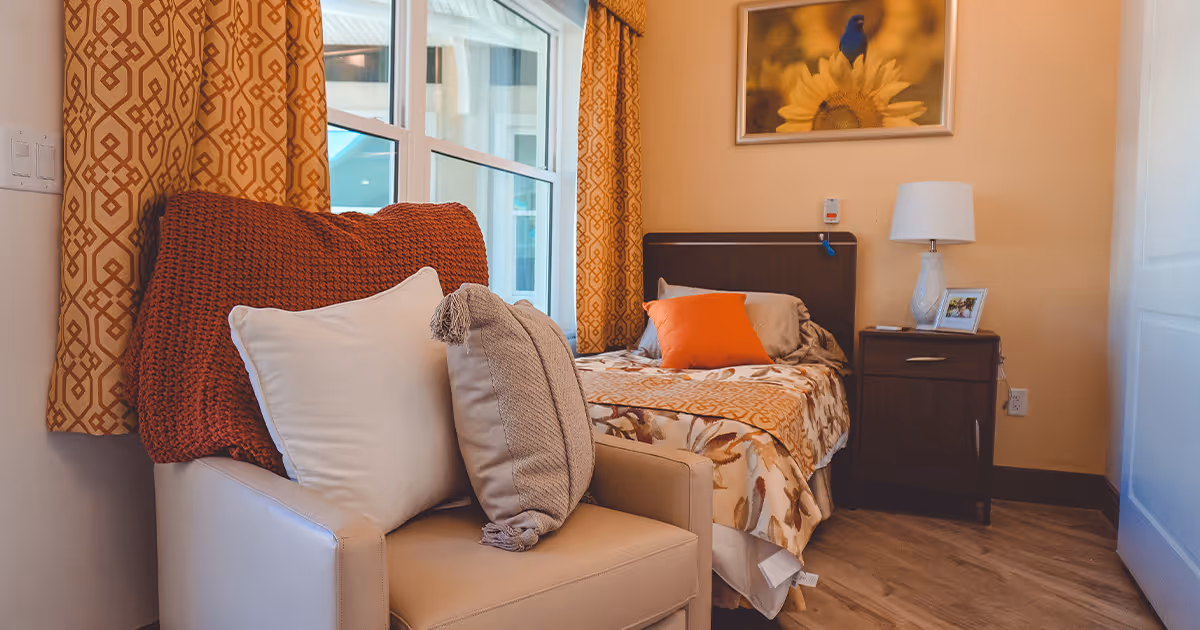 A cozy bedroom in an assisted living facility featuring a single bed with floral and orange bedding, an orange pillow, a wooden nightstand with a lamp and framed photo, a beige armchair with multiple cushions, patterned orange curtains, and a window letting in natural light. A framed picture of a blue bird on a sunflower hangs on the wall above the bed.