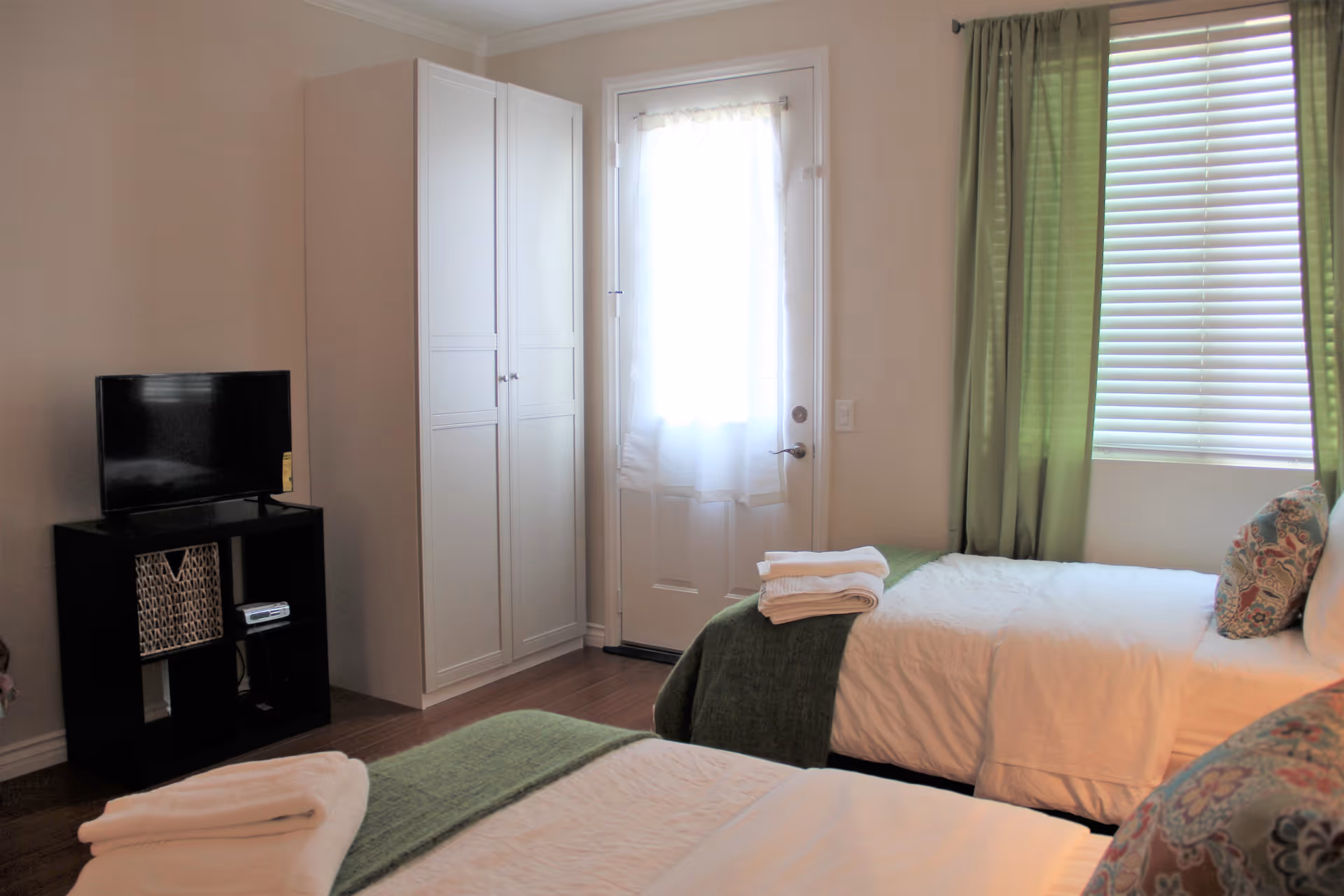 A bright bedroom with two single beds covered in white linens and green blankets, each with folded towels on top. There are decorative pillows on the beds. A window with green curtains and blinds is on the right wall. A white door with a sheer curtain is in the center background. A white wardrobe stands next to the door, and a black TV stand with a flat-screen TV and a basket is on the left side of the room.