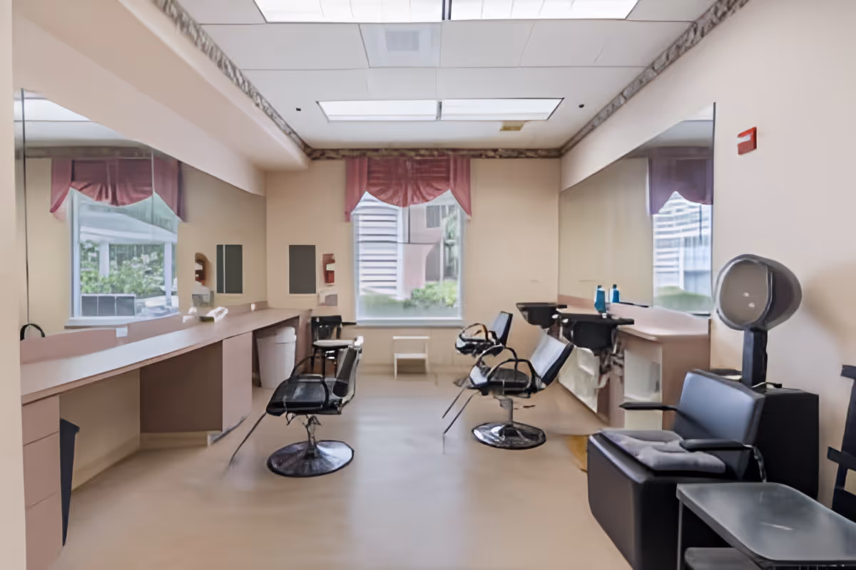 Interior of a salon area in a senior living facility with multiple black salon chairs, large mirrors on the walls, hair washing stations, and a window with pink curtains letting in natural light.
